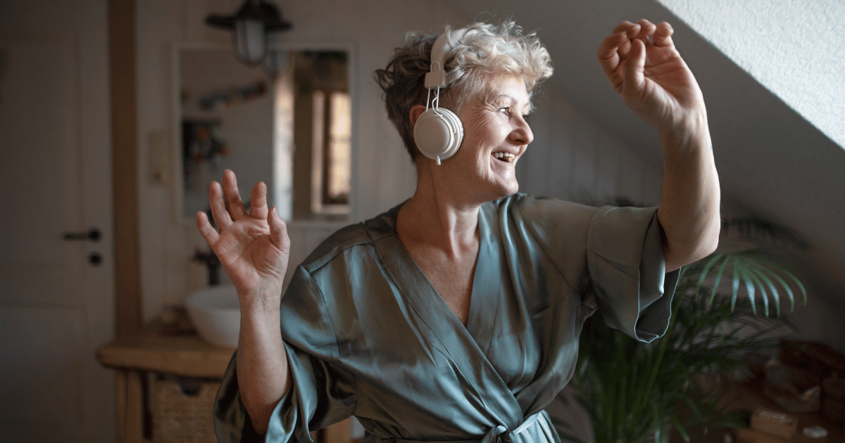 An older woman listening to headphones in her robe dancing with sunshine on her face from a nearby window